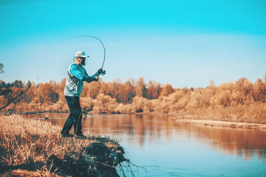 Angler casting a spinning rod from a grassy bank beside calm water, with autumn trees along the opposite shore under a blue sky.