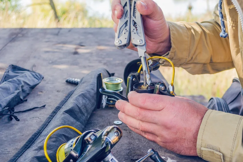 Close-up of an angler using a multitool on a spinning reel with its spool removed, with additional fishing equipment on the table.