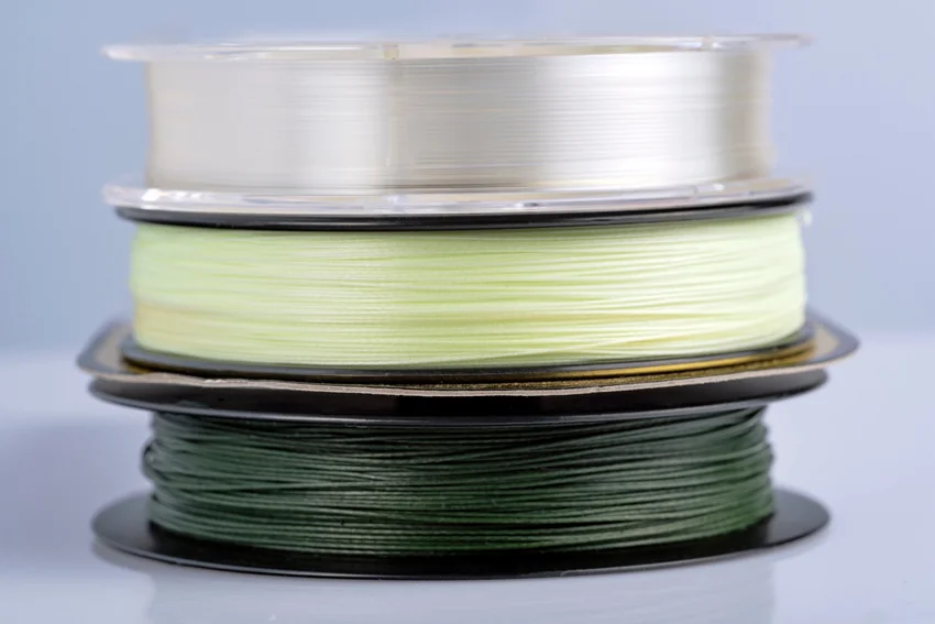 Three stacked fishing line spools containing clear, light green, and dark green line, arranged against a pale background with the strands visible.