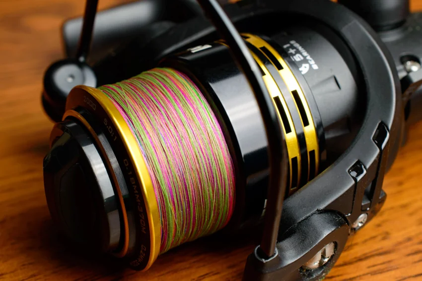 Close-up of multicolored fishing line wound around a black and gold spinning reel spool, with the reel resting on a wooden surface.