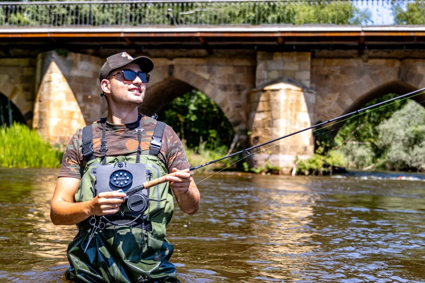 Smiling angler enjoying a sunny day while fishing in a river near a historic stone bridge, surrounded by lush green scenery.