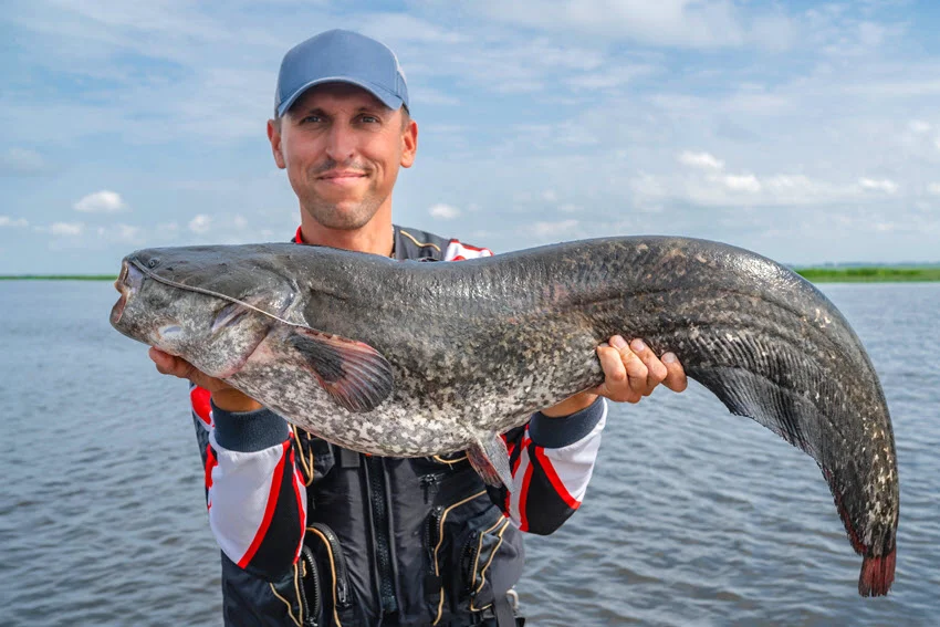 Happy fisherman proudly holding a big Catfish trophy on a boat, with fishing tackles nearby, capturing the excitement of a successful catch.