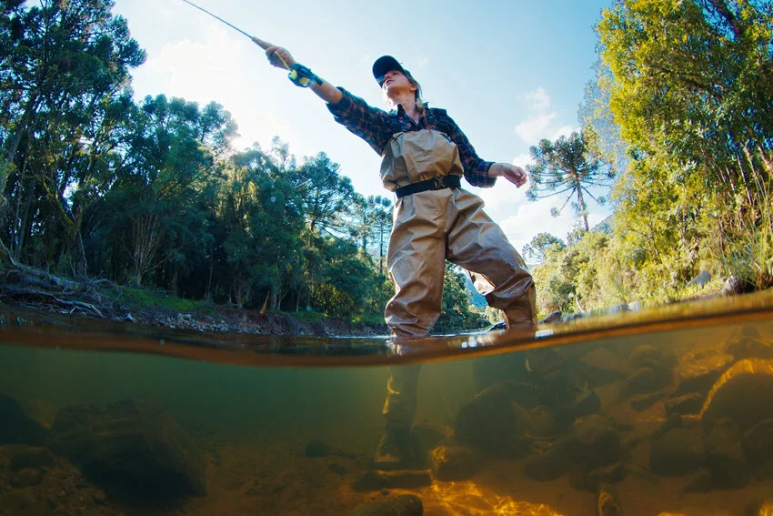 Woman angler standing knee-deep in a river, wearing waders and casting a fishing line into the flowing water during a peaceful fishing session.