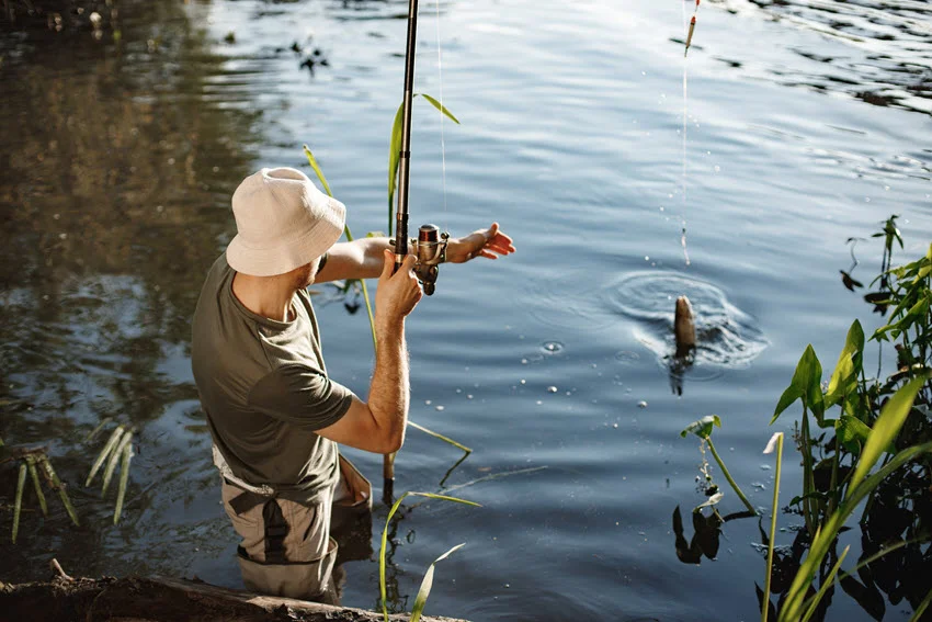 Back view of a young angler wearing a white hat and holding a fishing rod while reeling in a fish near the lake on a summer day.