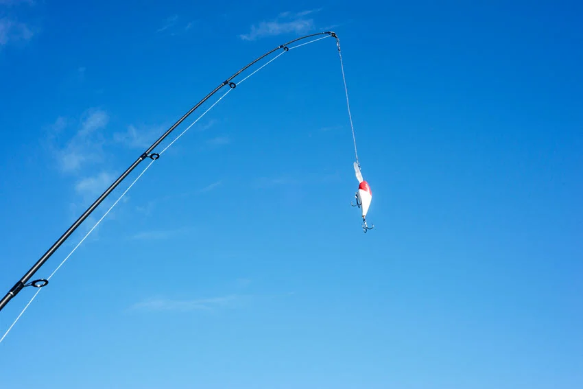 Spinning rod and brightly-colored crankbait against a blue cloudless sky, with fishing line cast and equipment on a cyan background.
