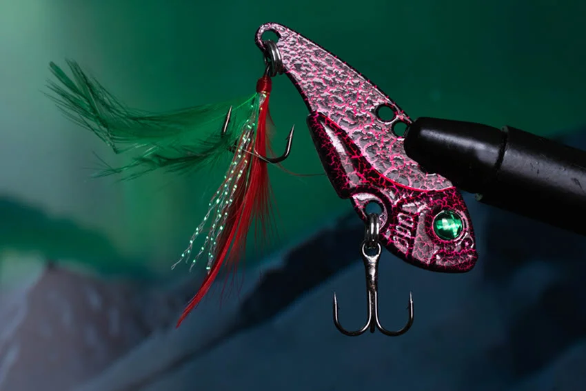 Detailed close-up view of a metallic crankbait lure with a textured pink body, green eye, and treble hooks, featuring red and green feather accents.