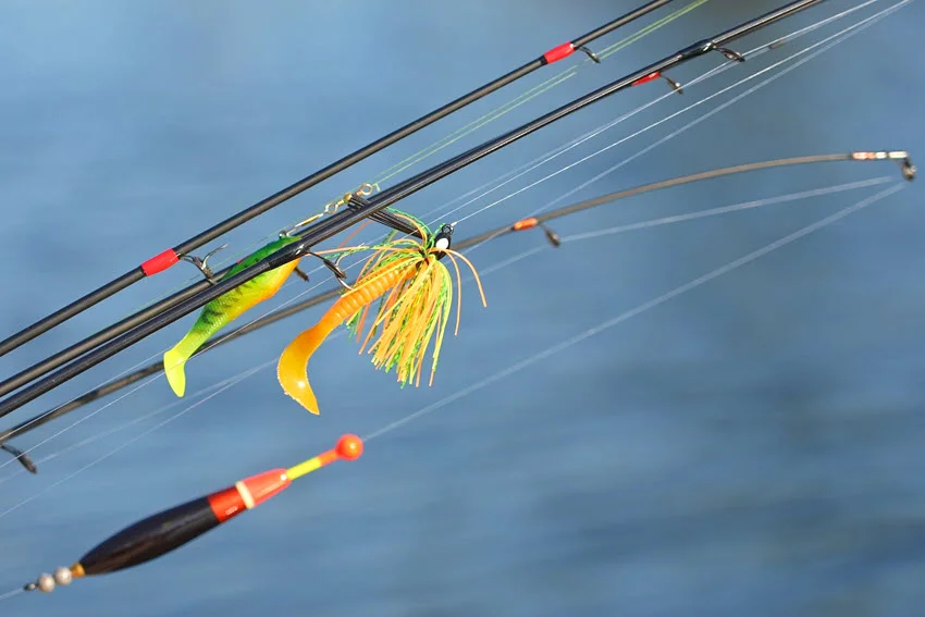 Fishing spinning and casting rods with tackles on a pier, focused on lines, bobber, and plastic jig with worm lures, with a blurred lake background.