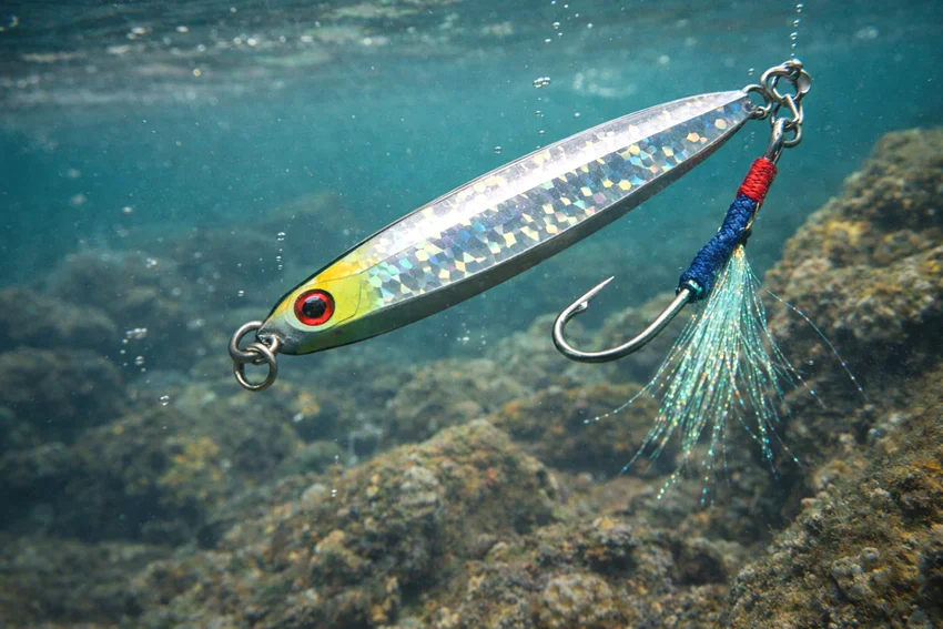 Diamond metal jig spoon suspended underwater above rocky Grouper structure, showing a vertical lure style used around reefs and deeper bottom habitat.