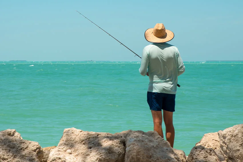 View of an angler casting from coastal rocks into clear turquoise water while searching for Jack Crevalle along the shoreline.