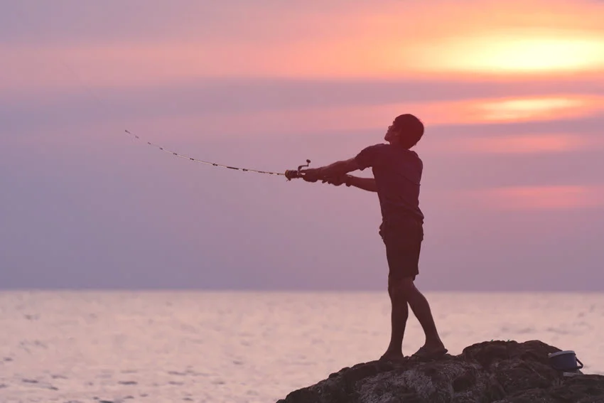 Action shot of an angler casting a lure from a rocky shoreline at sunset while targeting Jack Crevalle in coastal water.