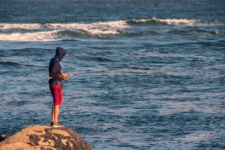 Angler jigging from a rocky shoreline in rough surf while targeting Jack Crevalle in fast-moving coastal water.