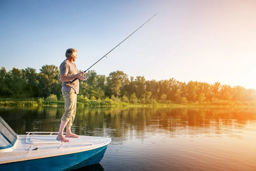 Side shot of a mature angler fishing from a motor boat at sunrise on calm water with a forested shoreline in the background.