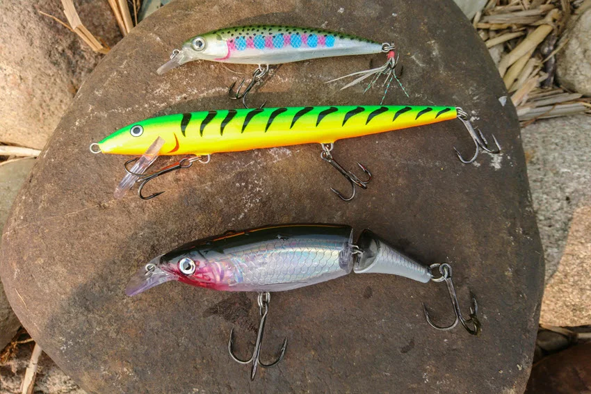Assortment of large jerkbait fishing lures with treble hooks, including Fire Tiger, X-Rap, and jointed designs, displayed on a rock surface.