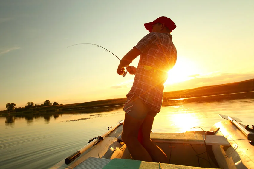 Young angler fishing from a small boat on a calm lake at sunset, with warm orange and pink colors reflecting on the water.