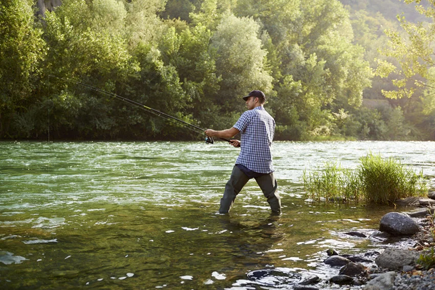Angler fishing from a shallow freshwater river bank, showing the type of shoreline cover where lure presentation and casting angles can matter for Peacock Bass.