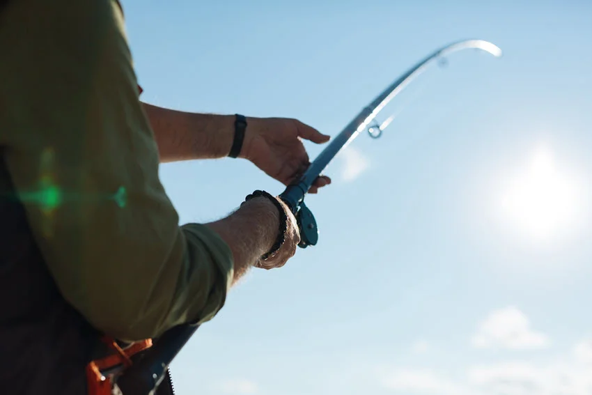 Man holding fishing tackle while fishing on a sunny morning, standing under a clear blue sky with bright sunlight illuminating the scene.