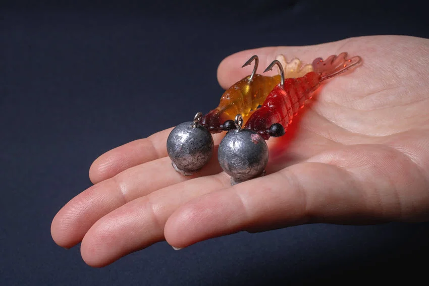 Two artificial shrimp-shaped fishing lures with jig heads in red and yellow, held by a white woman's hand.