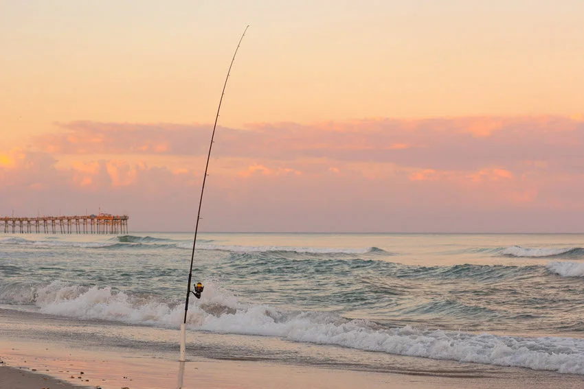 Fishing pole set in the surf on a beach with a pier in the background.