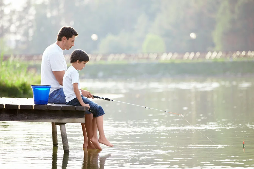 Father and son fishing from a wooden pier on calm freshwater, showing a simple beginner-friendly setup for learning how to cast and manage a rod.