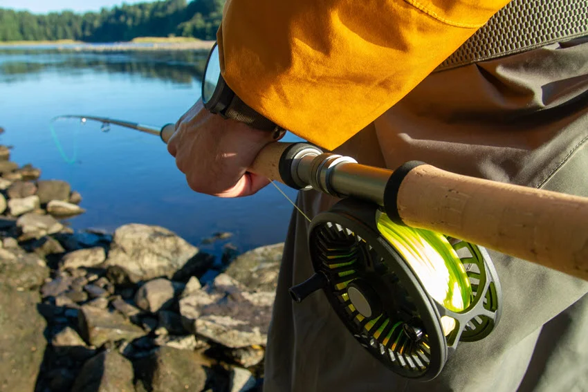 View of a fly fishing reel and rod near calm summer water, showing lightweight tackle used for Salmon fishing in clear river conditions.