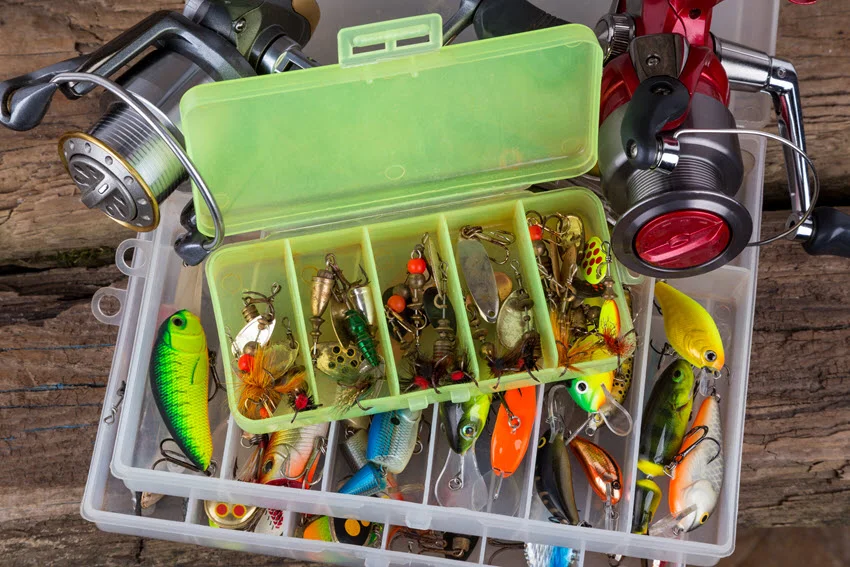 A top-down view of fishing tackle and colorful fishing baits neatly arranged in a box, set against a vertical wooden board background.