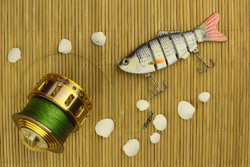 Top view of fishing reel, artificial small fish bait, shell bait presented on a bamboos mat.