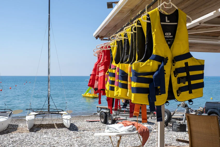Life jackets hanging on hangers, drying on a pebbled beach with sailboats and a beach visible in the background.