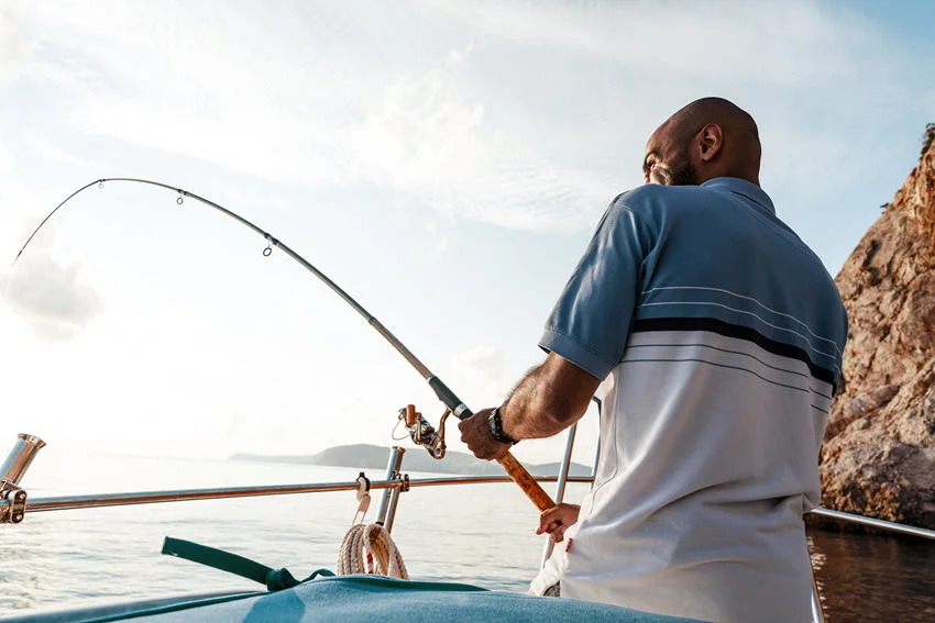 Close view of an angler standing on a sailboat while holding a fishing rod over calm blue offshore water.