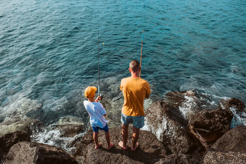 Father and son standing on rocks while fishing together near the sea on a sunny day.