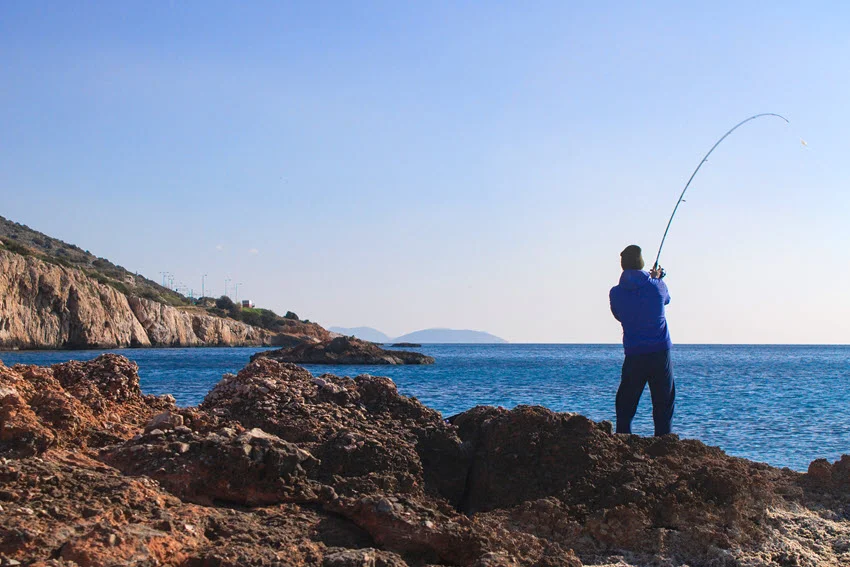 Fisherman standing on the shoreline with his rod bent under tension while fighting a fish.