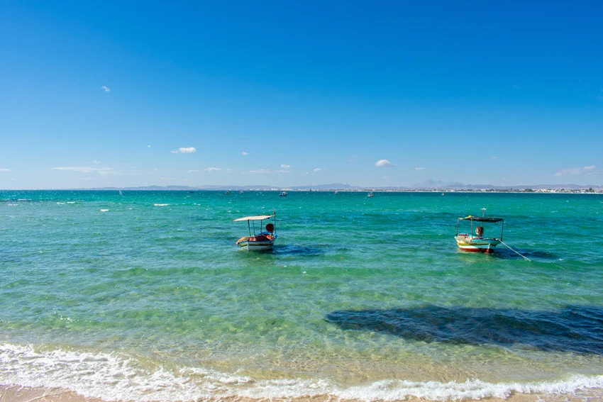 Two small oldy-style fishing boats drift calmly in a shallow turquoise ocean, very close to the sandy beach, on a sunny day, with a blue sky.