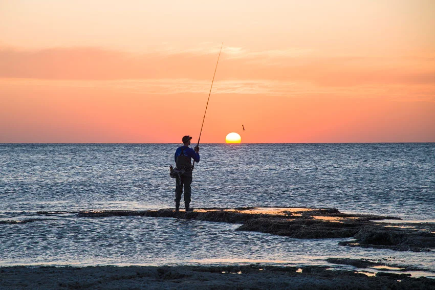 Angler casting from a rocky shoreline at sunset, showing the low light conditions when Snook often move shallow to feed.