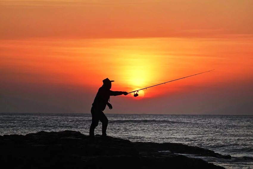 Silhouetted angler casting from coastal rocks at sunset, illustrating low-light shore fishing for Spanish Mackerel near the surface.
