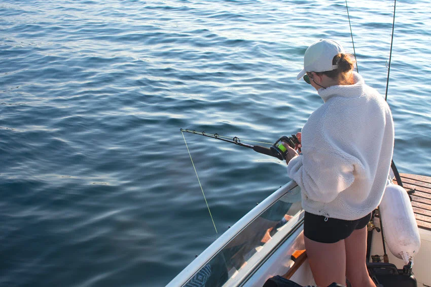 Female angler standing on a boat while jig fishing over calm blue ocean water.