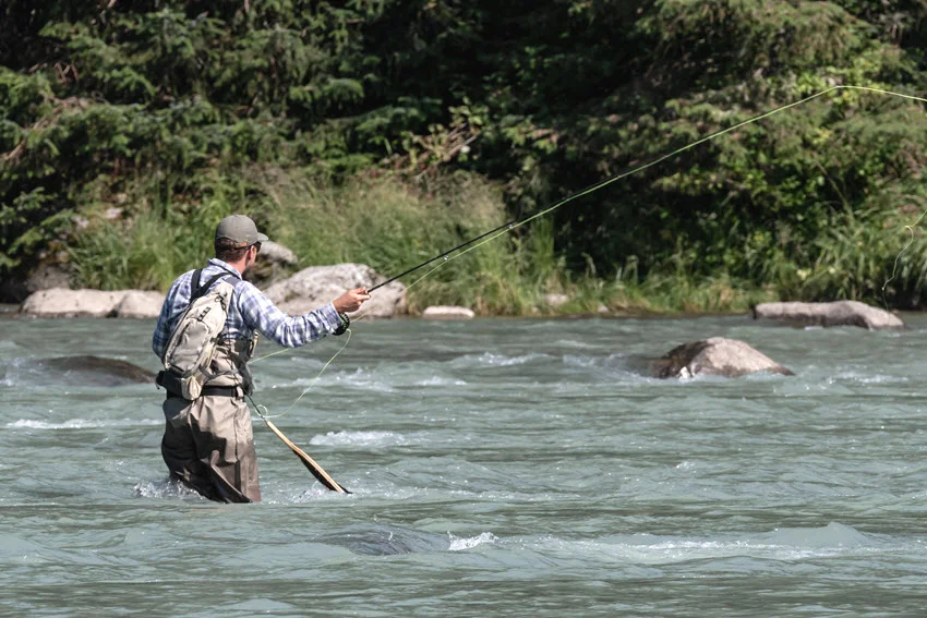 Back view of angler fly fishing in a river surrounded by forest, casting a line with a fly rod.