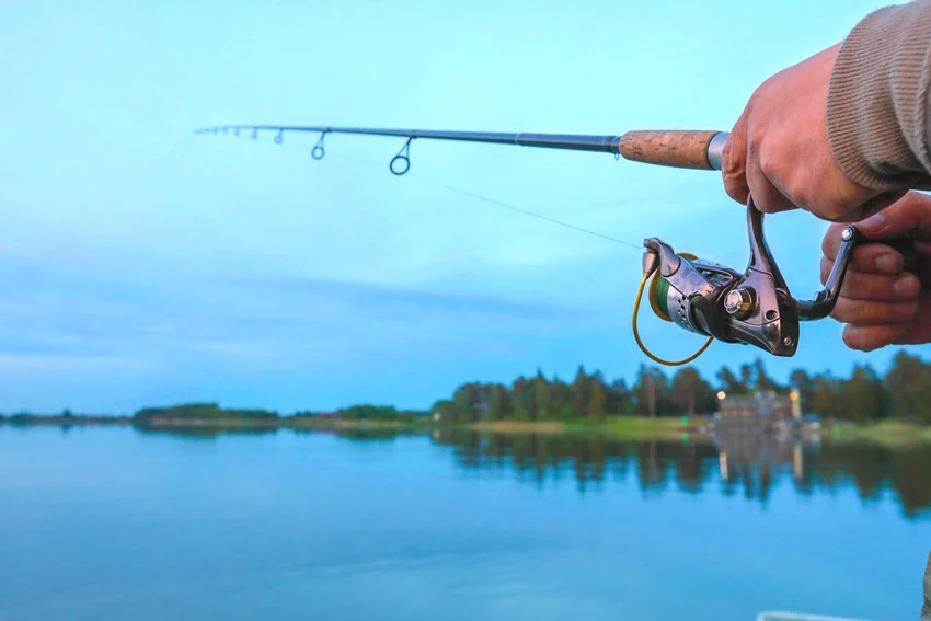Close-up view of a spinning fishing rod and reel, held by the angler, standing on a lake shore during sunset.
