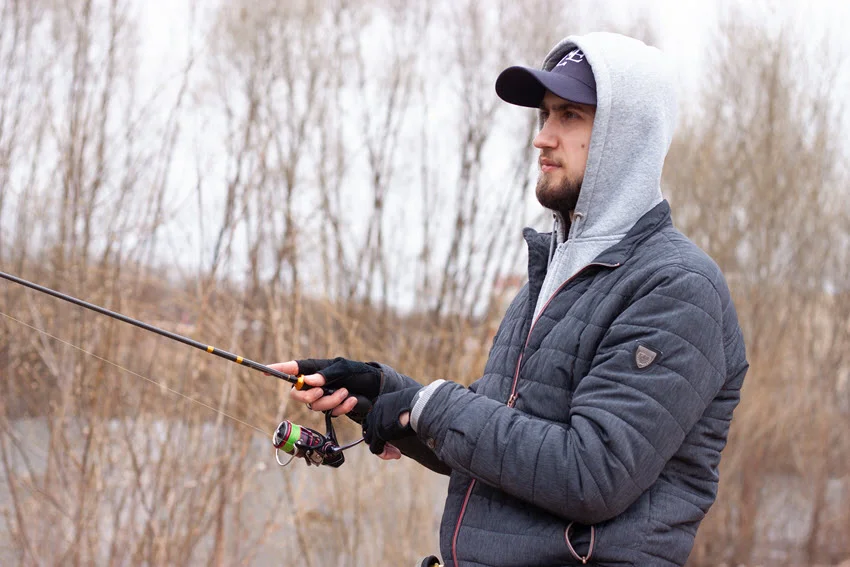 A young man with a beard holding a fishing rod wearing a light grey hood and dark blue jacket.