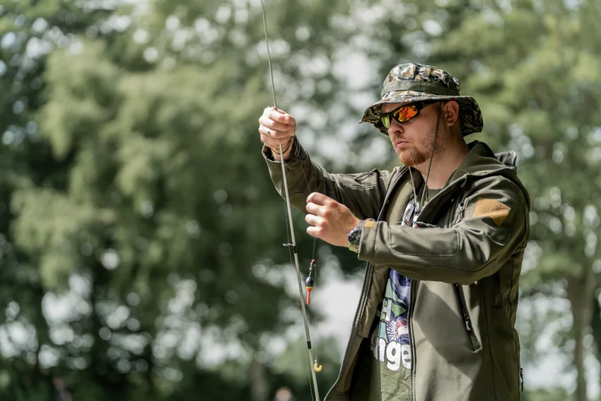 Angler wearing a hat and sunglasses while handling fishing line with both hands outdoors, with leafy trees softly blurred in the background.