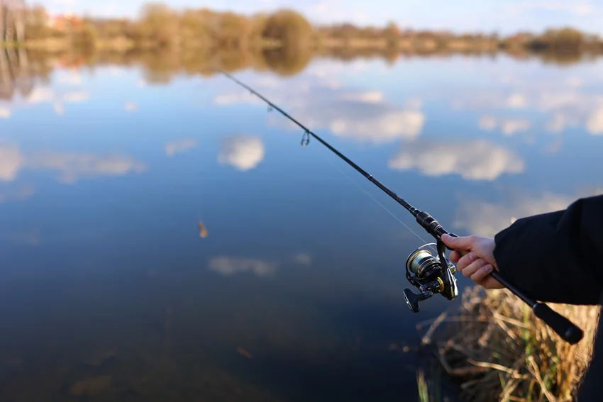 Angler holding a spinning rod and reel beside calm water, with reflected clouds and an autumn shoreline visible in the background.