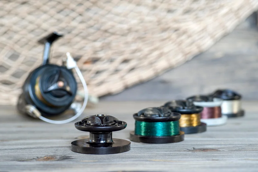 Several reel spools carrying different fishing line colors arranged on wooden boards, with a spinning reel and landing net in the background.