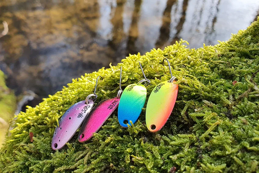 Assorted colorful spoon lures displayed beside a forest river used for Trout fishing.