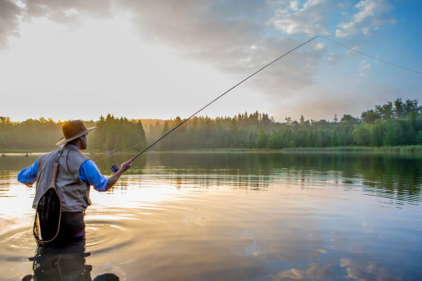 Young angler casting a fly rod across a peaceful forest river under soft evening light.
