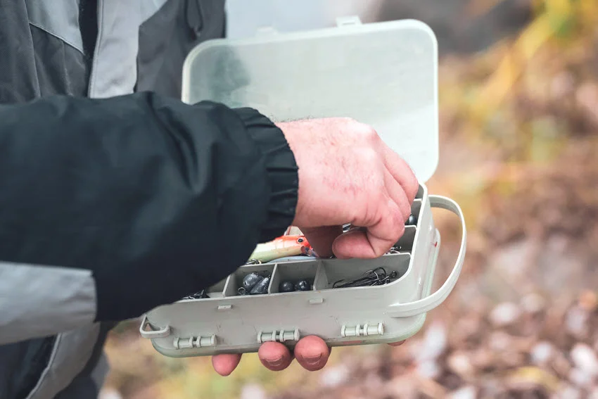 Focused view of an angler's hands holding a plastic tackle box filled with colorful lures and fishing hooks, with a blurry background suggesting preparation for a fishing trip.
