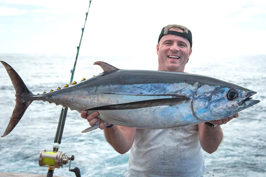 View of a happy angler posing with a freshly caught Blackfin Tuna in his hands, with fishing rods and the open ocean visible in the background.