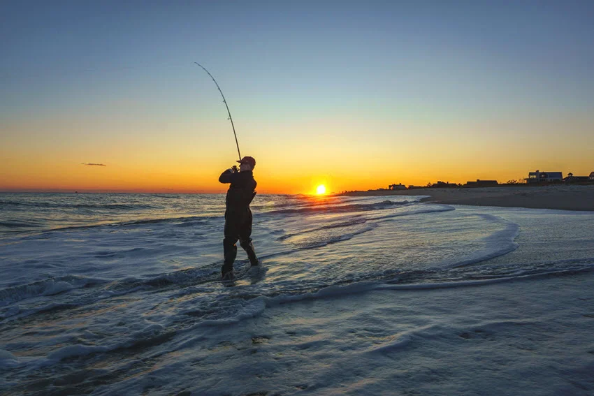 View of an angler fishing in ocean waves during sunset, with warm light reflecting off the water and silhouette framed by the horizon.