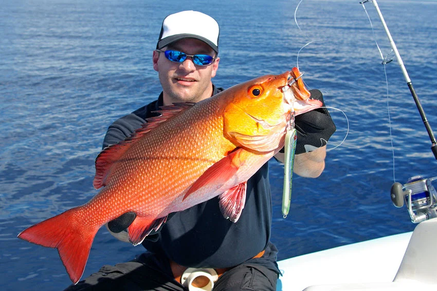 A happy, smiling angler with sunglasses sits on a boat, holding a Red Snapper.
