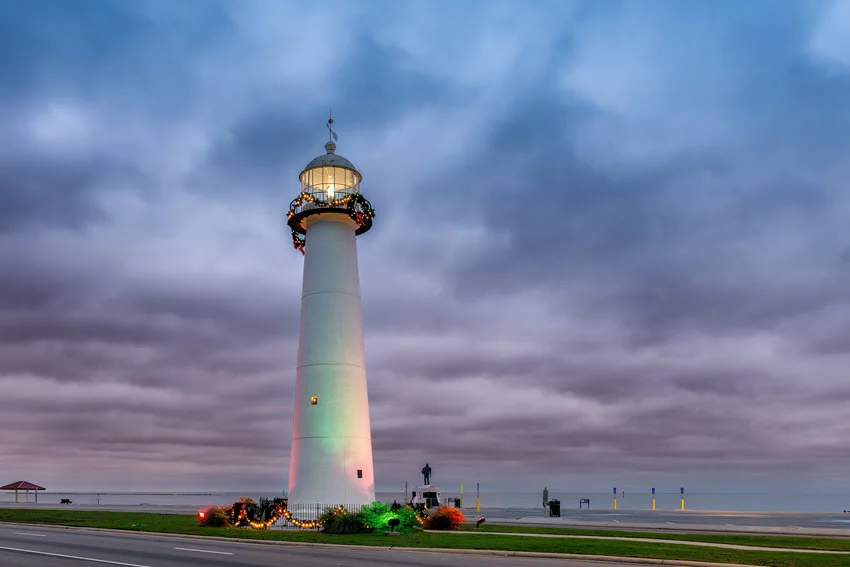 A beautiful white lighthouse in Biloxi Beach Bay stands illuminated by the vibrant sunset colors, towering against the backdrop of dramatic stormy clouds.