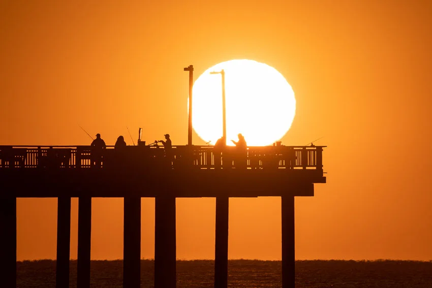 Magnificent sunset over a fishing pier, with a huge white sun disk and vibrant orange sky, as several anglers stand silhouetted while fishing.