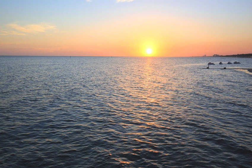 View of a stunning sunset above Biloxi Bay waters, with warm colors reflecting off the calm surface and silhouetted shoreline in the distance.