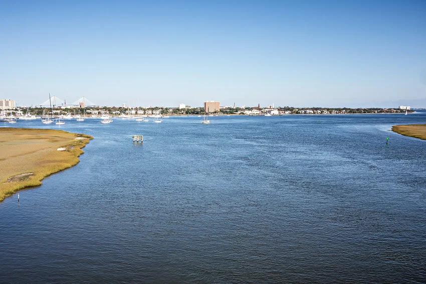 Charleston, South Carolina, skyline across a broad river, with sailboats, marsh-lined banks, and a distant bridge beneath a clear blue sky.